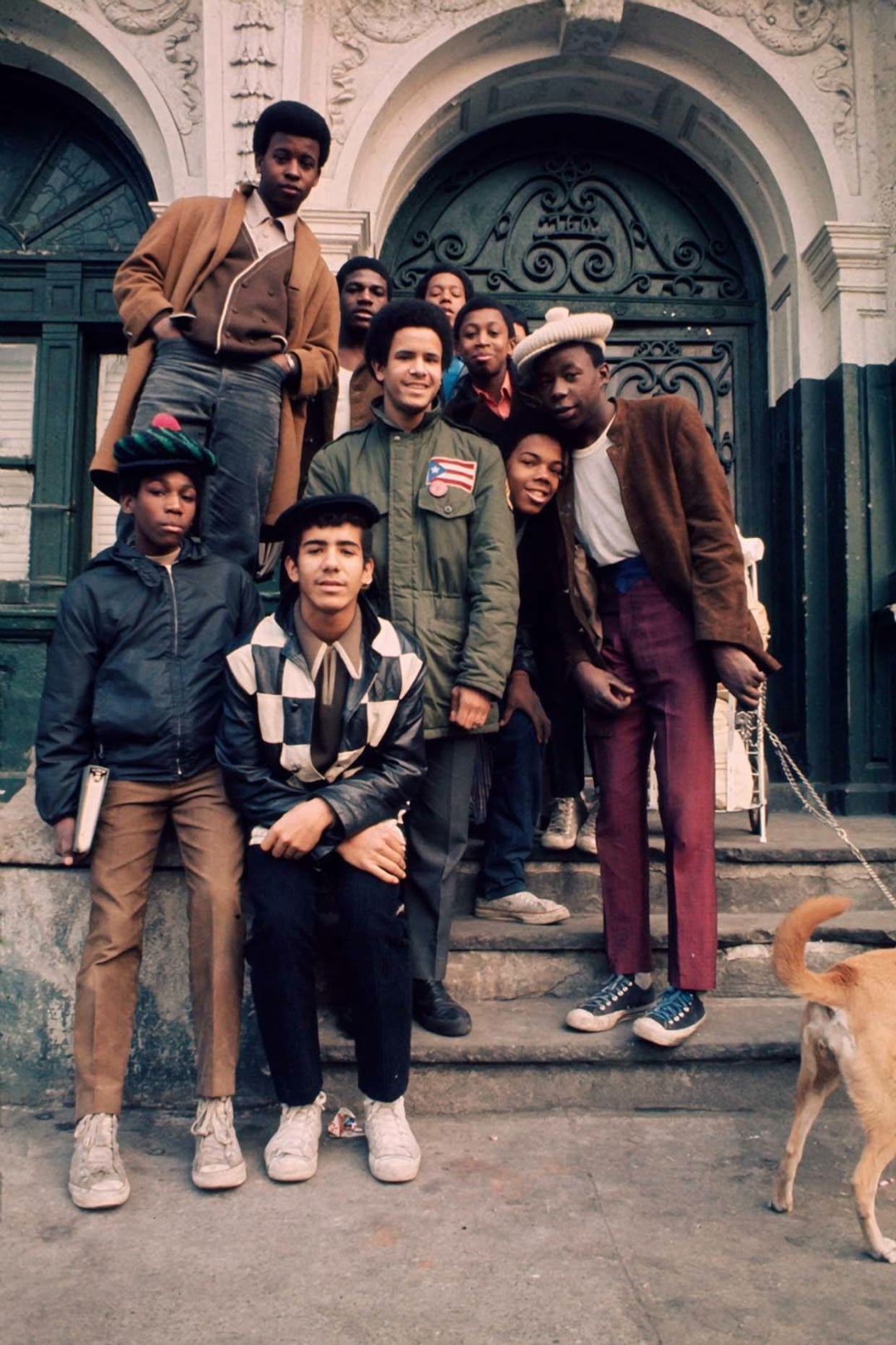 A group of eight boys pose on stone steps in front of an ornate building entrance. One holds a book, another holds a dog on a leash. Most are dressed in jackets and hats, and they all look toward the camera, appearing relaxed and friendly.