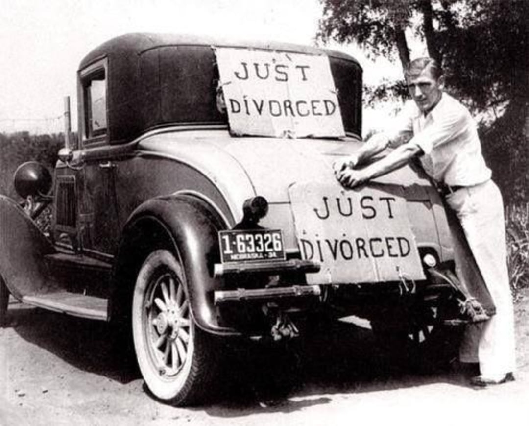 A man attaches a sign reading "JUST DIVORCED" to the back of a vintage car, which already has another identical sign on the spare tire. The car has a 1932 Nebraska license plate.