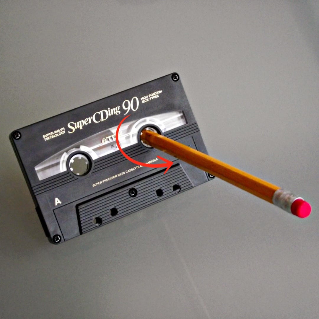 A pencil is inserted into the reel of a cassette tape, illustrating how to manually rewind or tighten the tape. An arrow shows the direction to turn the pencil.