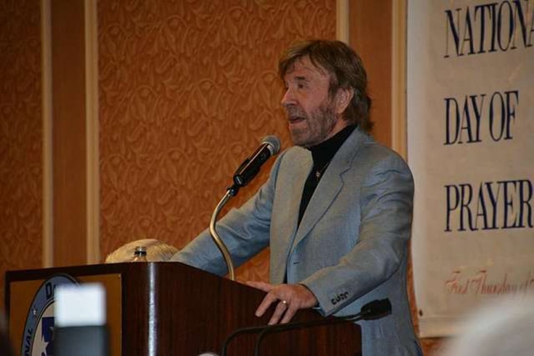 A man in a gray suit speaks into a microphone at a podium during an event. A large sign in the background reads "National Day of Prayer." The room has ornate wallpaper.
