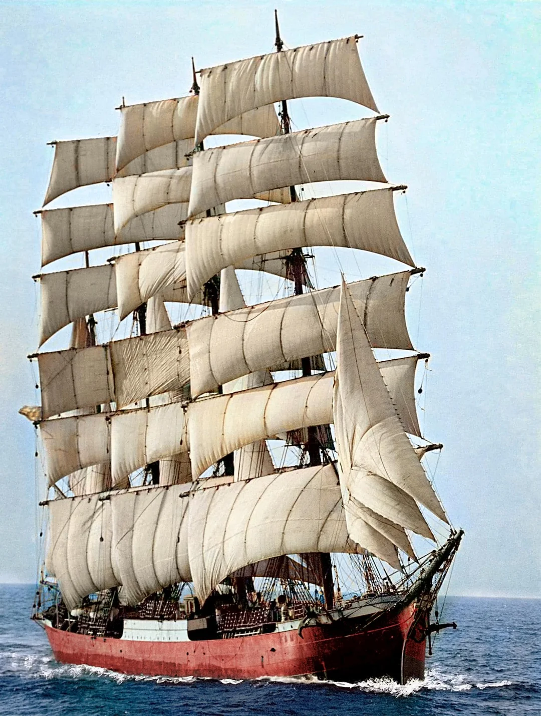 A large, red-hulled sailing ship with multiple tall masts and white sails fully unfurled glides across a calm blue sea under a clear sky.