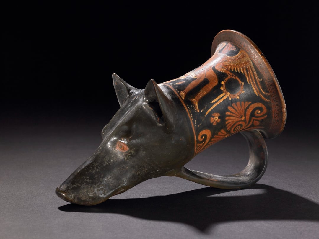 Ancient Greek ceramic drinking vessel shaped like a black dog’s head with a decorated flared neck featuring painted figures and patterns, lying on its side against a dark background.
