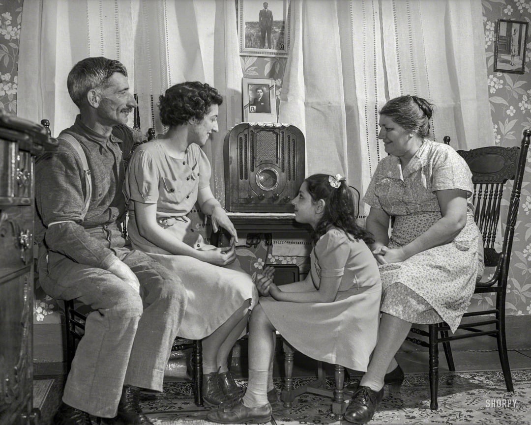 A family of four, two adults and two girls, sit together in front of a vintage radio in a cozy, patterned room, listening attentively. Curtains, framed photos, and floral wallpaper create a warm, nostalgic atmosphere.