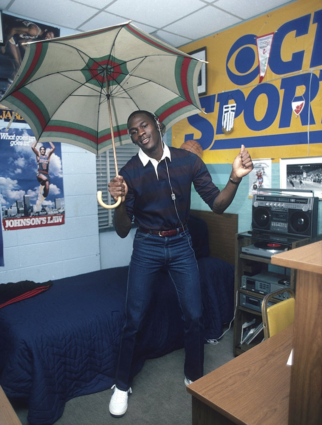 A young man stands in a dorm room, smiling and holding an open umbrella indoors. He wears headphones, a dark shirt, blue jeans, and white sneakers. The room has sports posters, a bed, and a stereo on a desk.