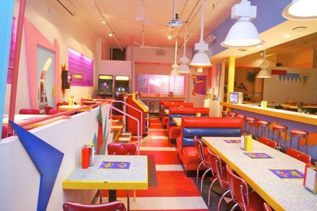 A brightly colored retro diner with red chairs, booths, checkered floors, pastel walls, and neon lighting. Condiment bottles sit on tables, and arcade machines are visible in the background.