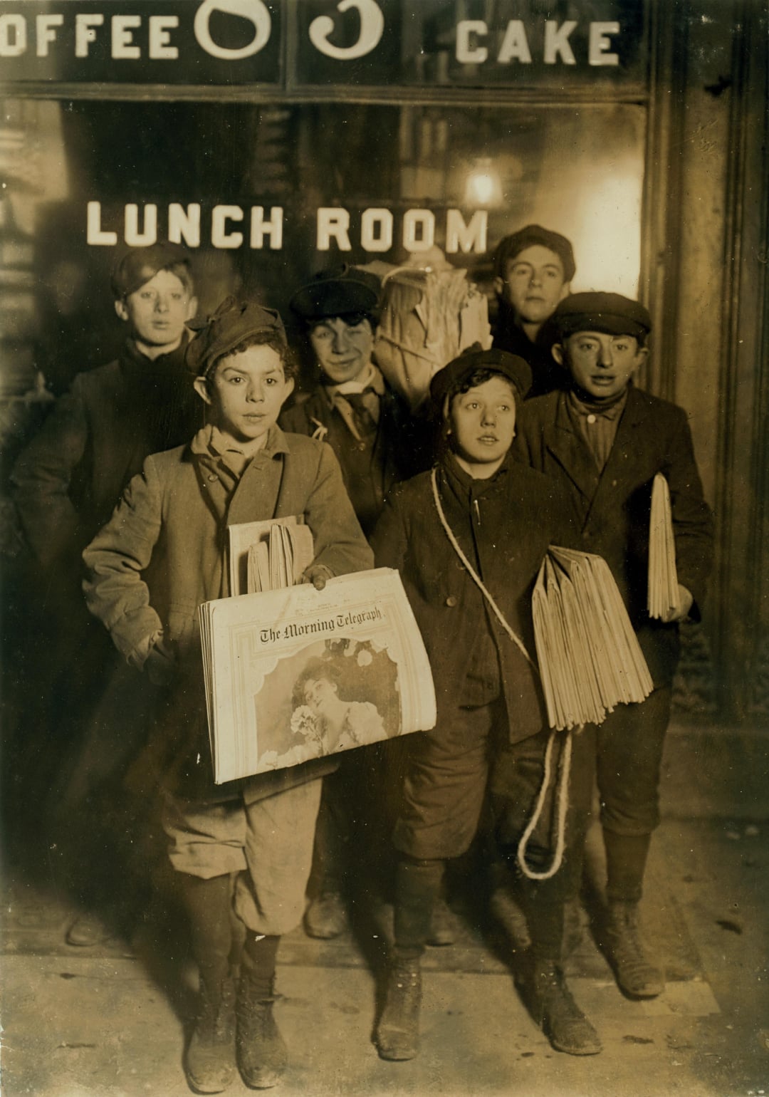 A group of six boys wearing caps and coats stand outside a building, holding newspapers for sale. The sign behind them reads “COFFEE 5¢ CAKE” and “LUNCH ROOM.” The scene appears to be from the early 1900s.