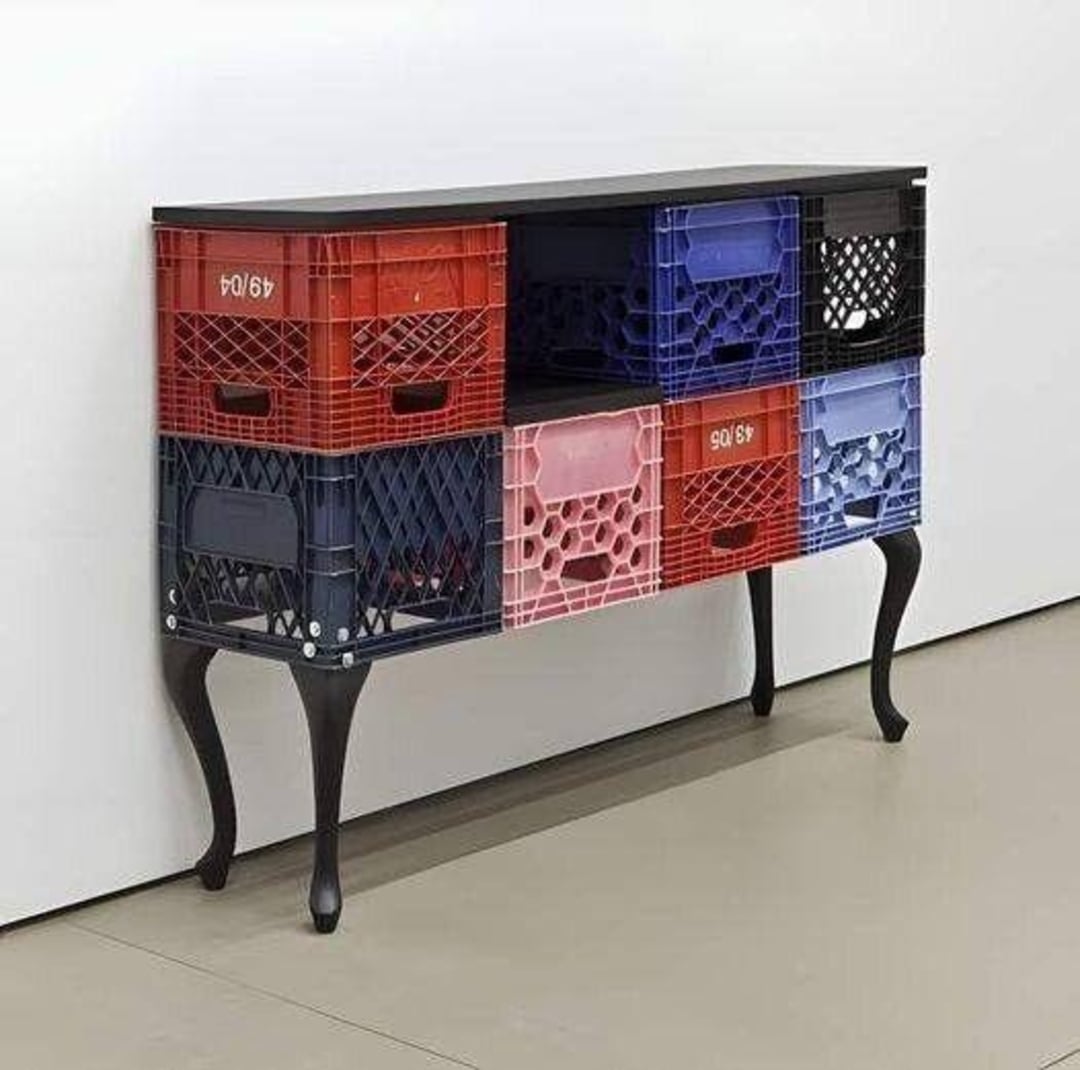 A console table with black ornate legs, featuring colorful plastic milk crates as drawers, arranged in two rows under a black tabletop, standing against a white wall.