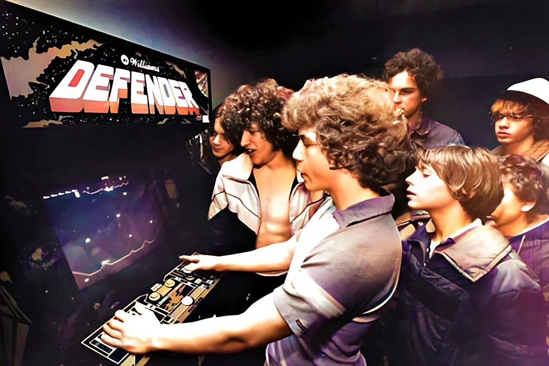 A group of teenagers in vintage clothing gather around a Defender arcade game machine, watching intently as one of them plays. The scene has a nostalgic 1980s atmosphere.