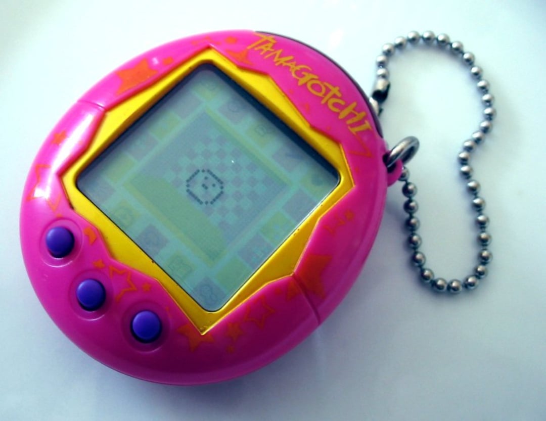 A close-up of a pink Tamagotchi virtual pet device with a chain keyring, yellow trim, three purple buttons, and a pixelated character on its screen.