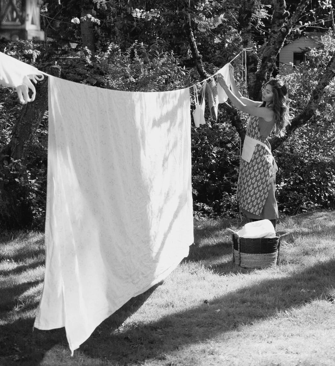 A woman hangs laundry on a clothesline outdoors in a sunlit garden, with a large white sheet and other clothes drying, and a basket of laundry beside her.