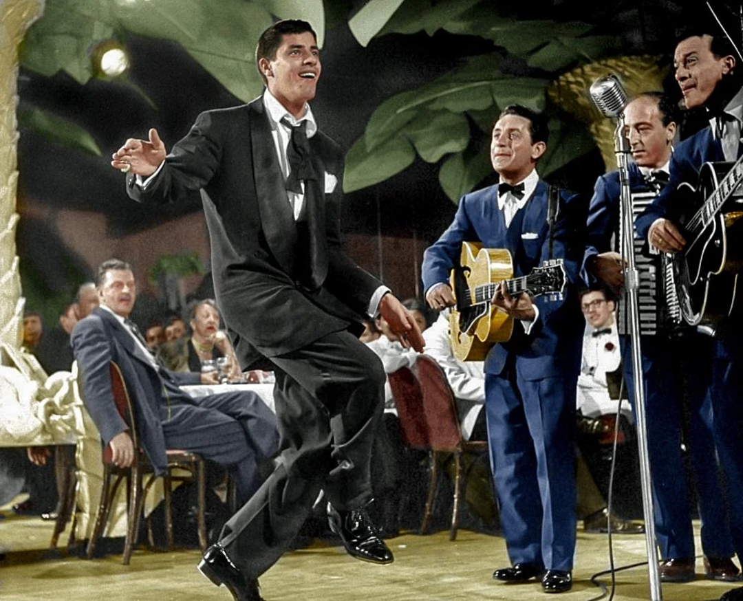 A man in a tuxedo dances energetically on stage beside a jazz band playing guitar and accordion, with an audience seated at tables in the background in a lively club setting.