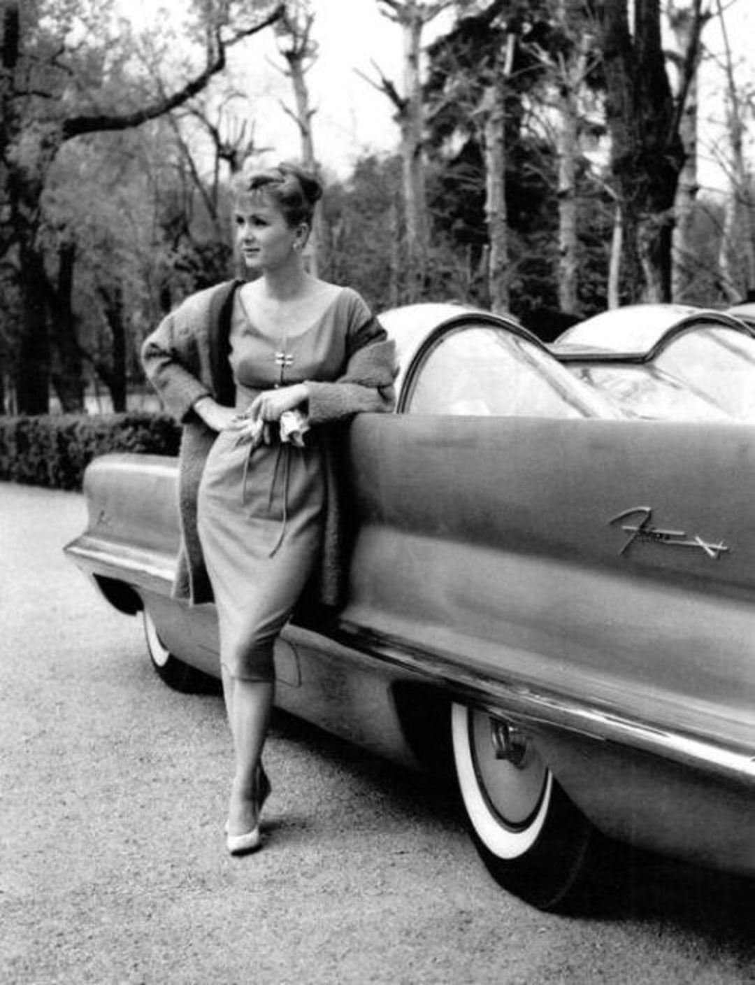 A woman in a dress and cardigan leans against a sleek vintage car with whitewall tires, parked on a tree-lined road. The car has curved glass panels and the name "Firebird" visible on its side.