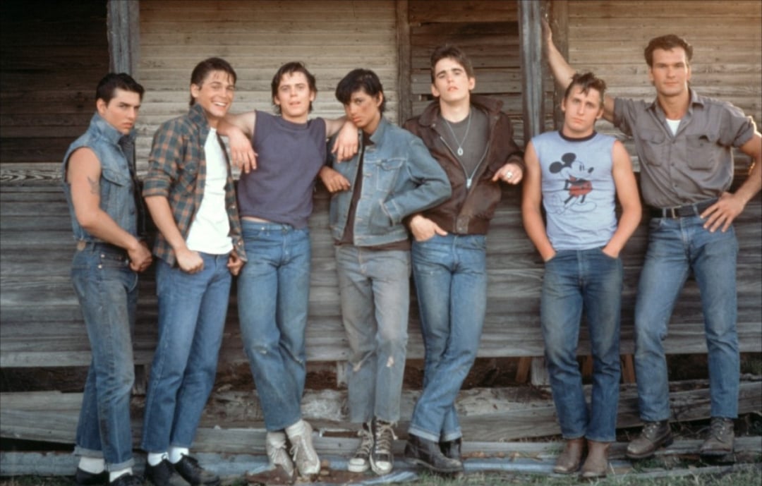 Seven teenage boys in casual 1960s-style clothing stand side by side in front of a weathered wooden building, posing together and looking at the camera. Most wear jeans and t-shirts or denim jackets.
