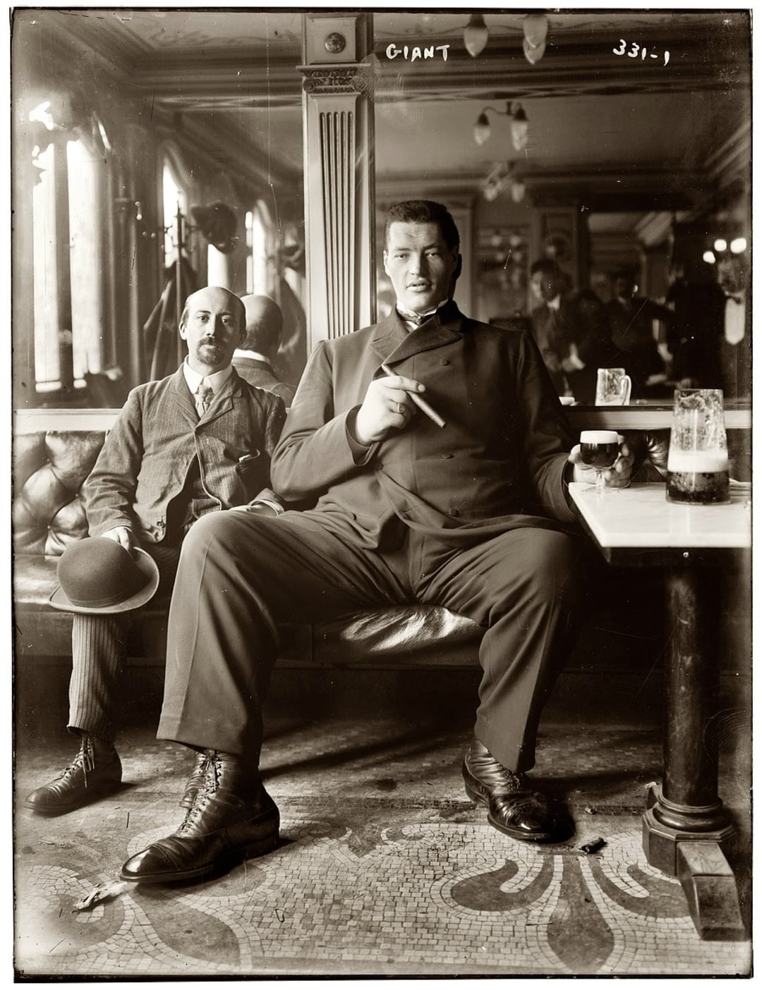 Two men sit in a vintage bar; one is extremely tall and large, holding a cigar, while the other, much smaller man holds a hat. The table has drinks on it, and their reflections appear in a mirror behind them.