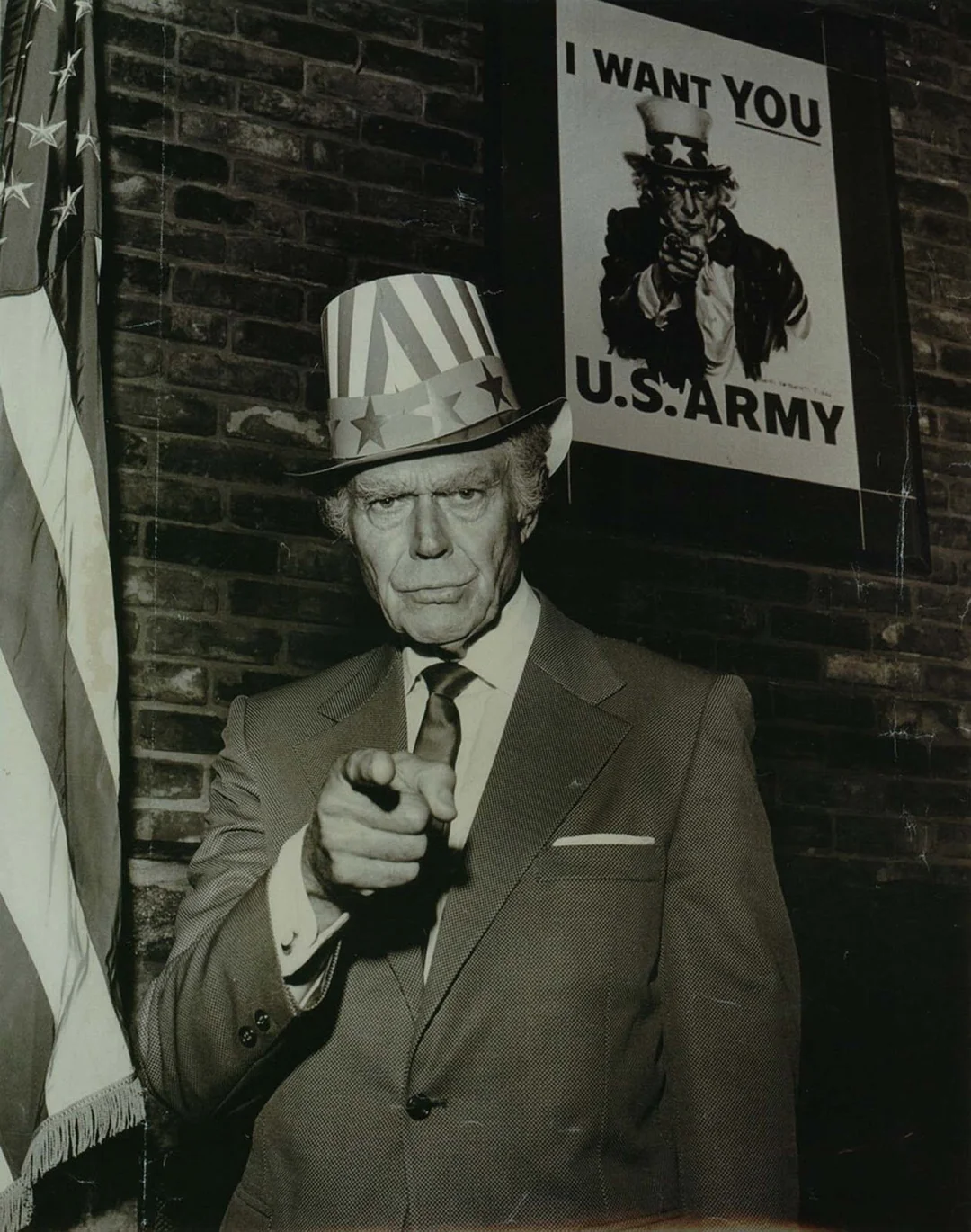 An older man in a suit and a stars-and-stripes Uncle Sam hat points at the camera, standing next to a U.S. flag and beneath a "I Want You U.S. Army" Uncle Sam recruitment poster.