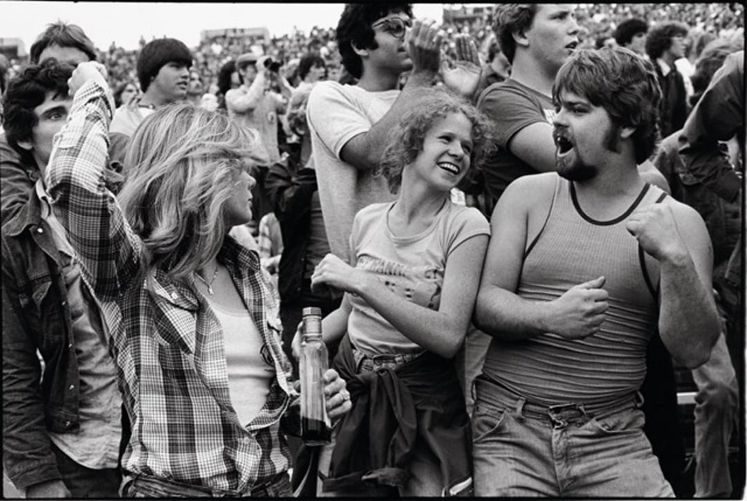 A group of young adults cheer and dance energetically in a crowded outdoor setting. One woman holds a bottle, while others smile and raise their fists in excitement, suggesting a lively concert or festival atmosphere.