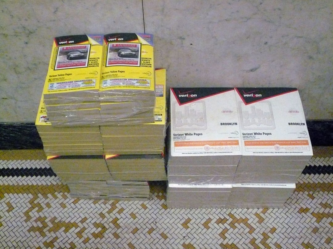 Two stacks of phone books sit on a tiled floor against a marble wall. The left stack has yellow Verizon directories, while the right stack has white Verizon Brooklyn White Pages phone books.