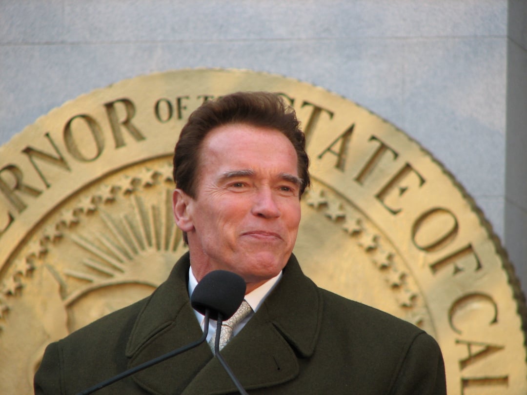 A man in a dark coat stands in front of a large gold seal reading "Governor of the State of California," speaking into a microphone.