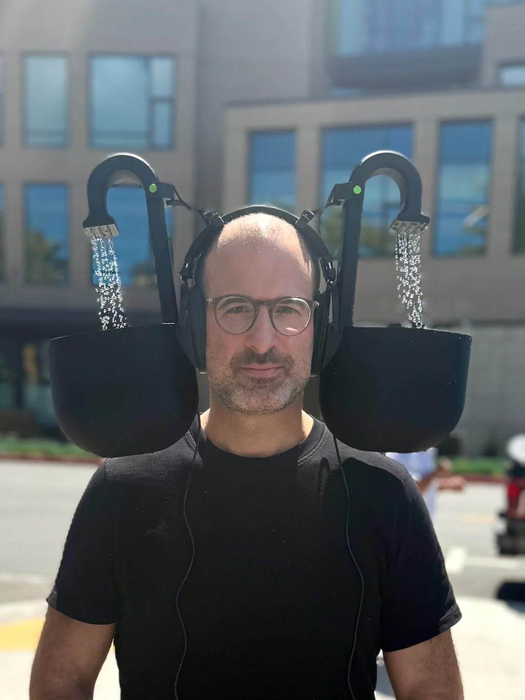A man wearing glasses and a black t-shirt stands outside, wearing headphones with attached devices that pour water into large cups hovering near his ears. A modern building is visible in the background.