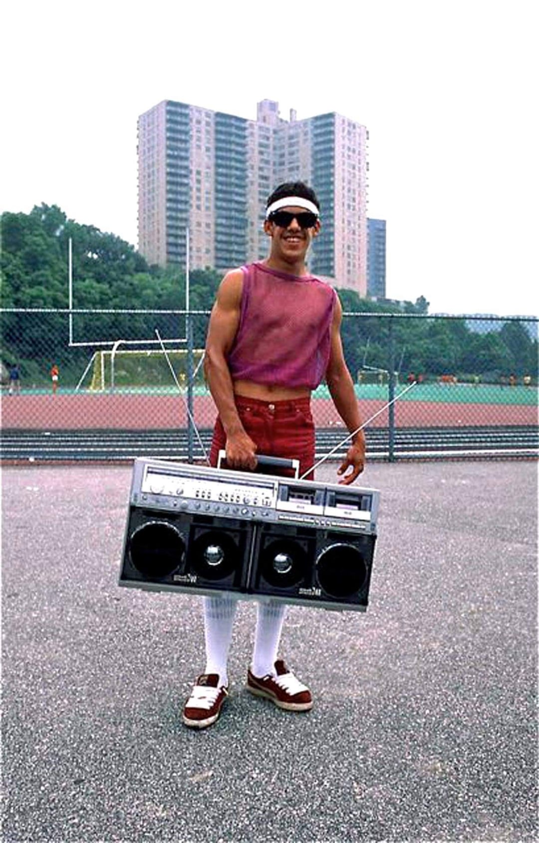 A man in athletic shorts, a sleeveless mesh shirt, sunglasses, and headband stands on pavement holding a large boombox. In the background are a chain-link fence, sports field, trees, and tall apartment buildings.