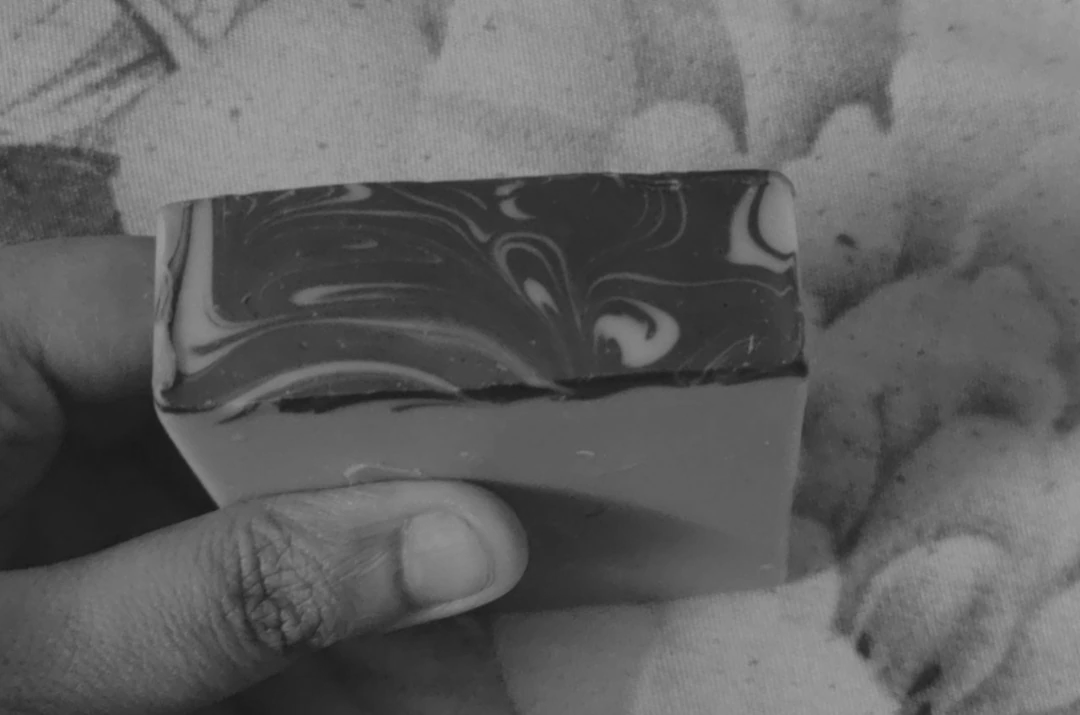 A hand holding a rectangular bar of soap with a marbled, swirled pattern on the top surface, placed against a patterned background. The image is in black and white.