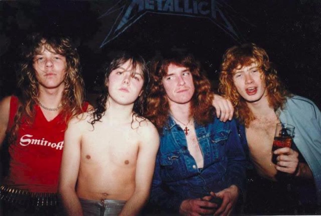 Four young men with long hair pose together in front of a Metallica sign; two are shirtless, one wears a red tank top, and one wears an open denim shirt while holding a drink.