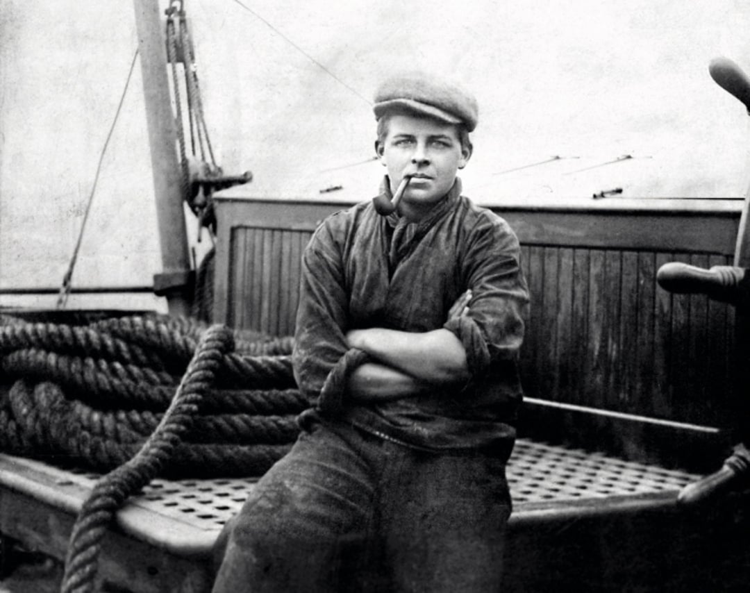 Young person sits on a wooden boat deck with arms crossed, wearing a flat cap and smoking a pipe. Thick coiled ropes and wooden ship structures are visible in the background. The image is black and white.
