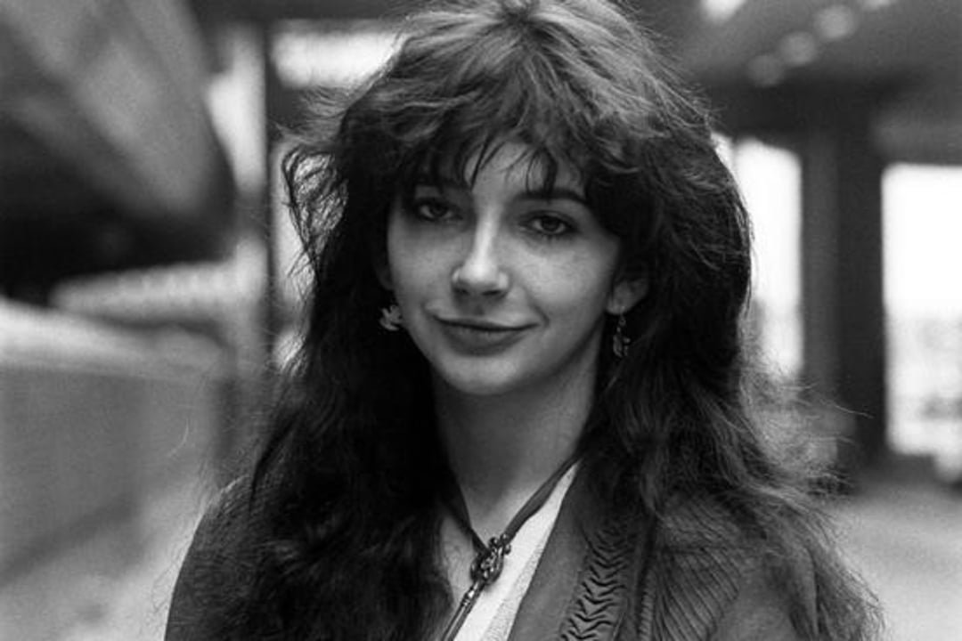 A young woman with long, wavy dark hair smiles softly at the camera. She is wearing a necklace and earrings, and stands outdoors in an urban setting. The photo is black and white.