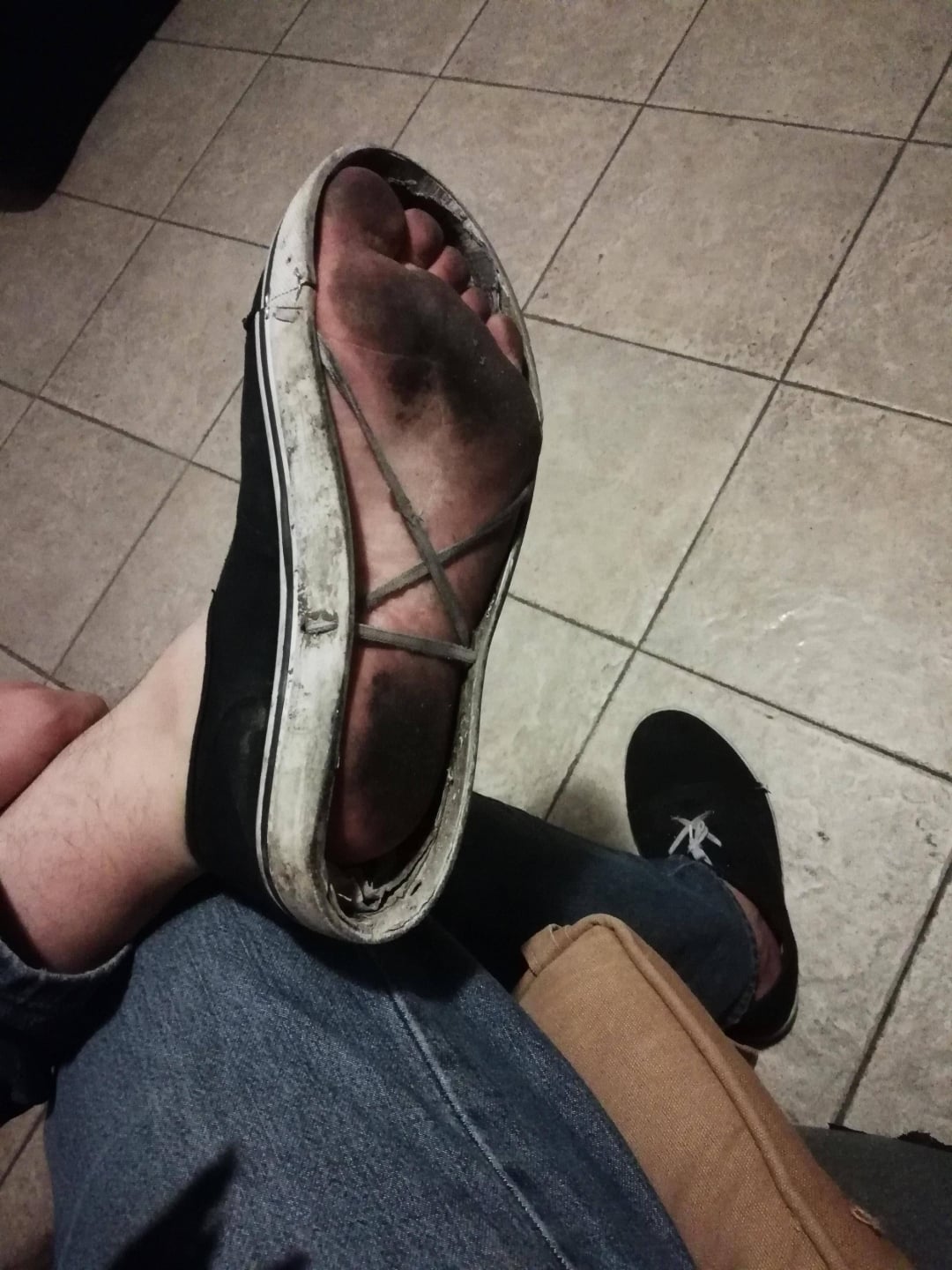 A person wearing worn black and white sandals with a very dirty sole shows the bottom of their foot. They are sitting on a tiled floor, wearing jeans, and another black shoe is visible on their other foot.