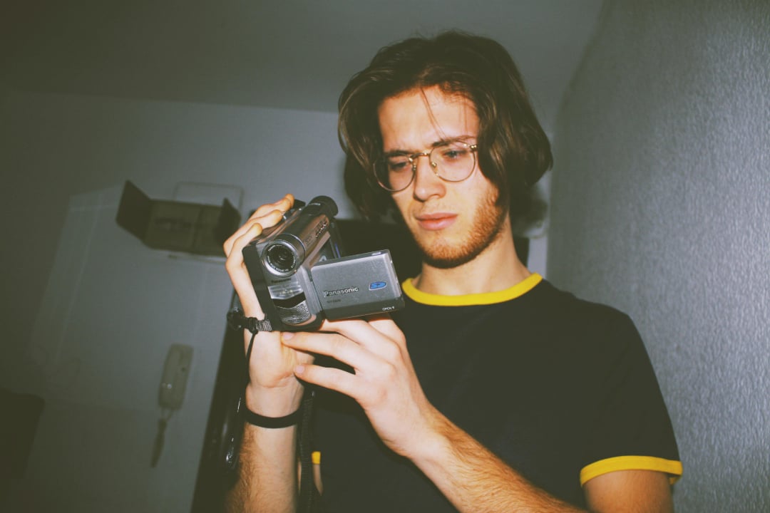 A person with long hair and glasses, wearing a black t-shirt with yellow trim, is looking down while holding and examining a handheld video camera indoors.
