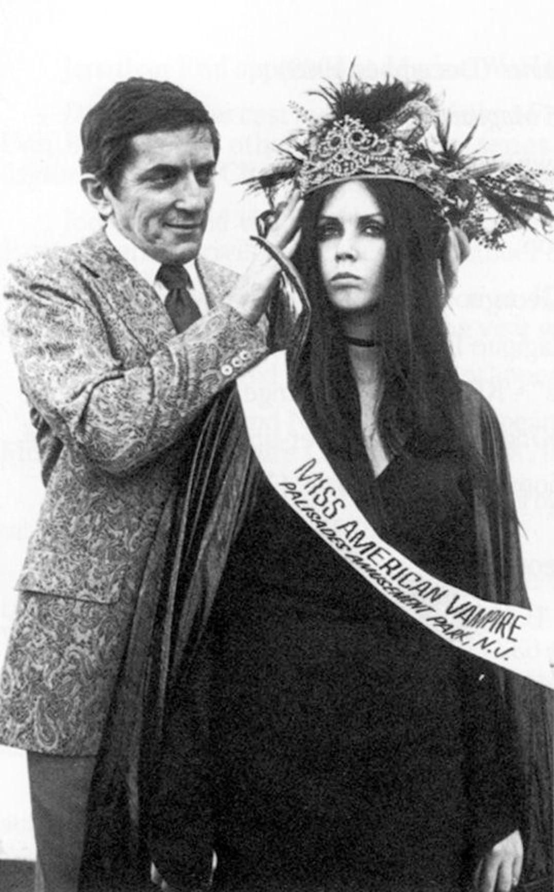 A man in a patterned suit places a crown on a young woman wearing a dark cape, choker necklace, and a sash reading "Miss American Vampire." Both have serious expressions in this black-and-white photo.