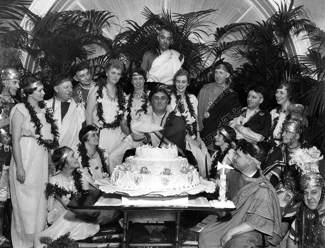 A group of people in Roman-style costumes gather around a large decorated cake, with one person seated at the center and others standing or sitting nearby, smiling and celebrating under a decorative arch and leafy plants.