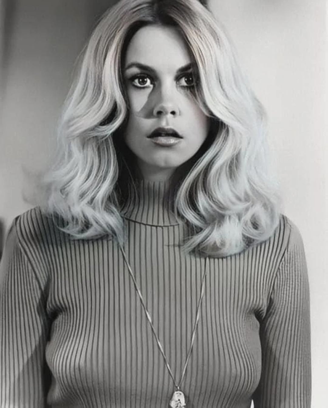 A woman with voluminous, wavy blonde hair wears a ribbed, high-neck sweater and a long necklace. She looks forward with a surprised expression in this black and white photo.