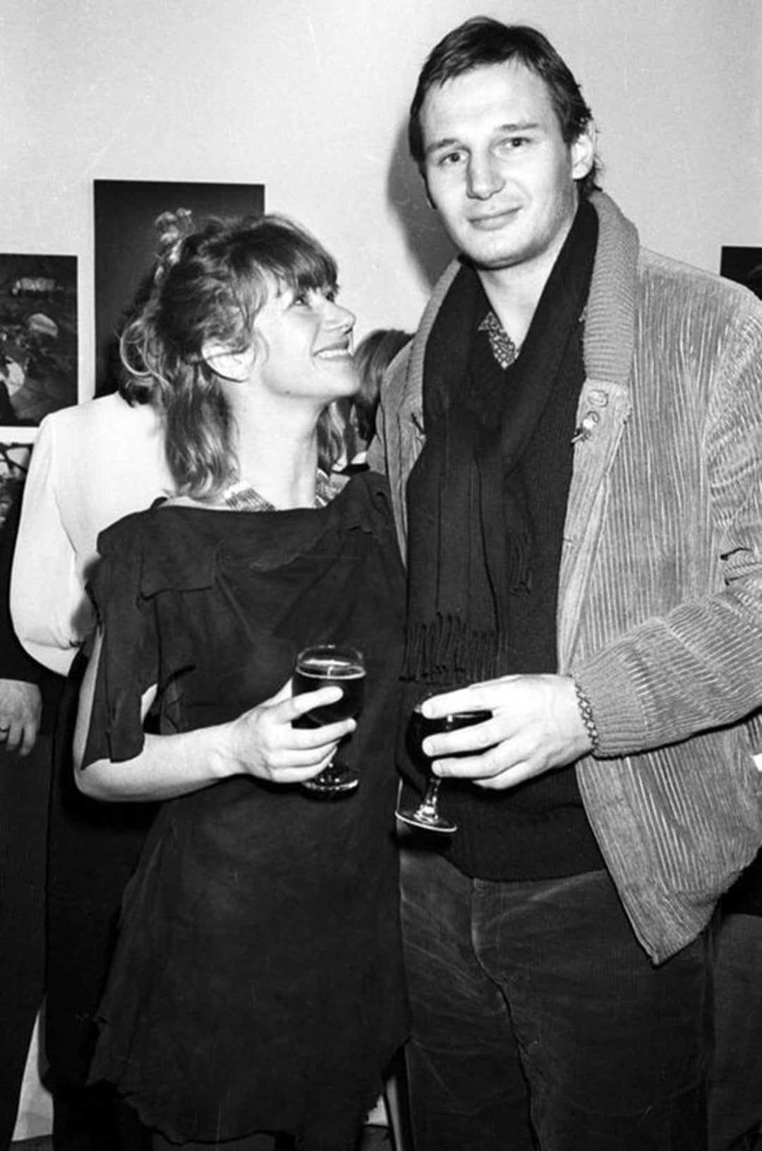 A woman smiling up at a man while both hold drinks at a social gathering. The man wears a jacket and scarf; the woman wears a loose dress. They are standing close together with people and artwork in the background. The photo is in black and white.