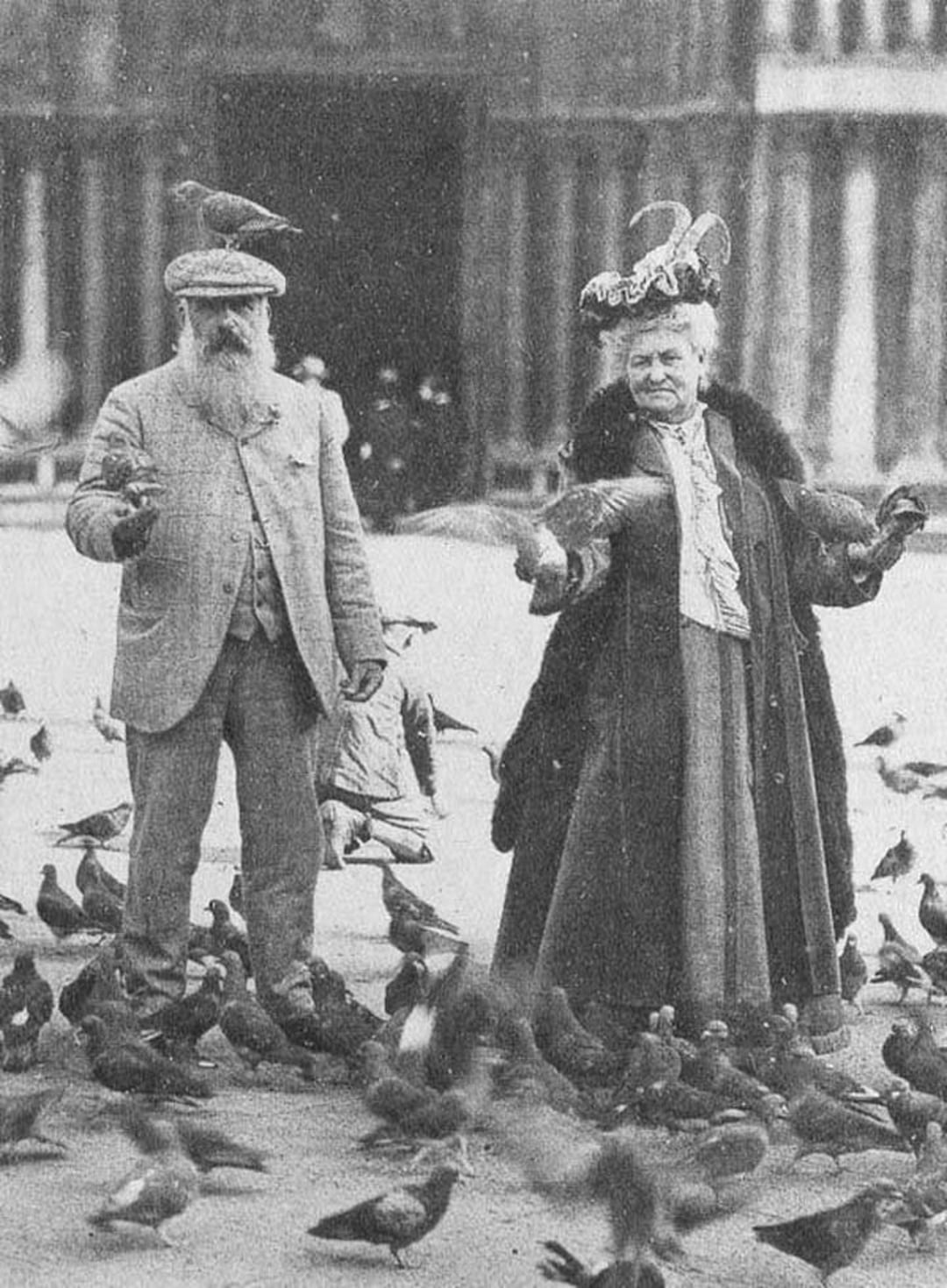 An old black-and-white photo of a man and a woman feeding pigeons outdoors. The man has a beard and wears a hat and suit; the woman wears a long dress, coat, and decorated hat. Numerous pigeons surround them, with one on the man's head.