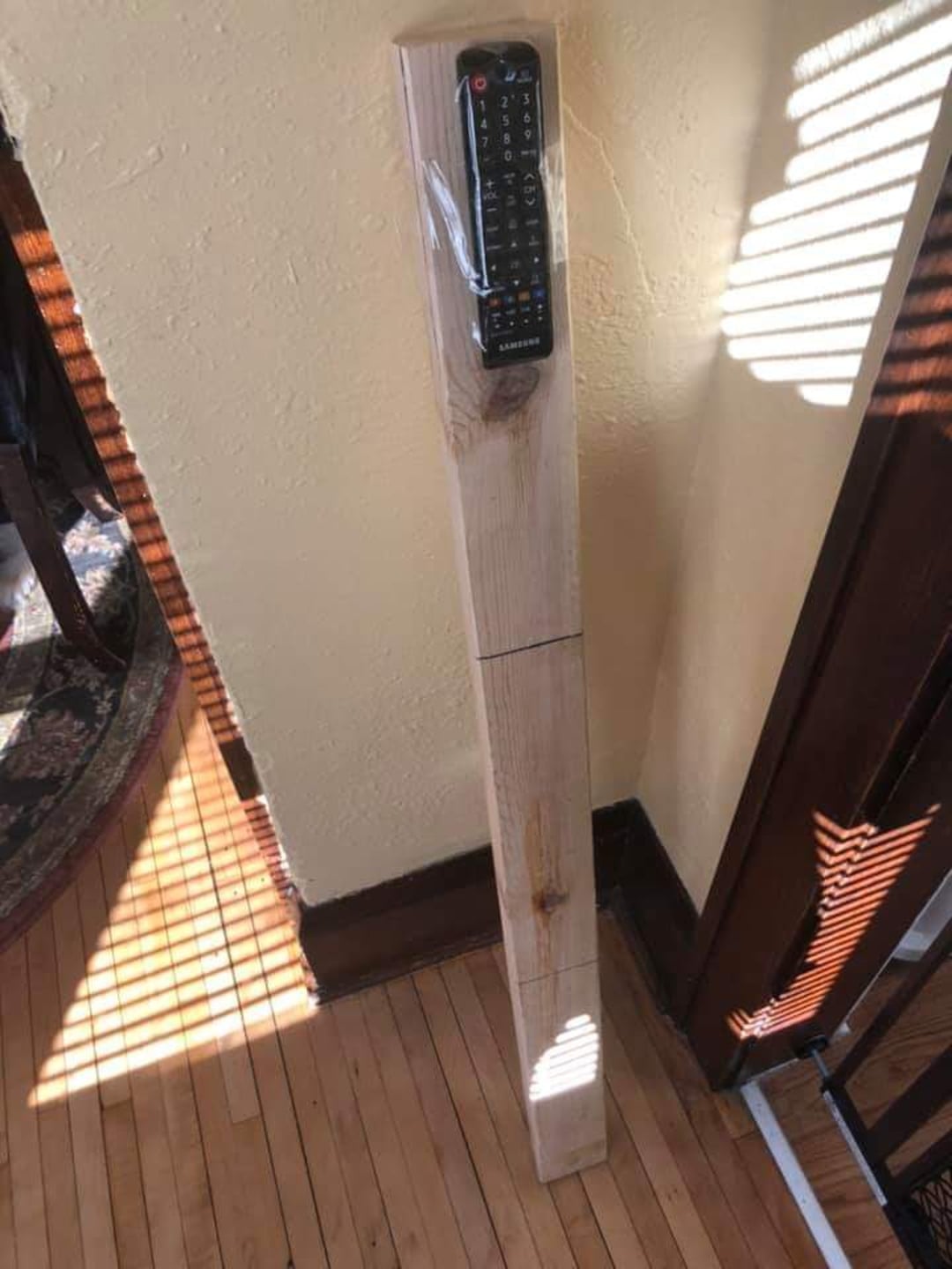 A TV remote control is taped to the top of a long wooden plank leaning against a wall in a sunlit room with wooden floors and shadows from window blinds.