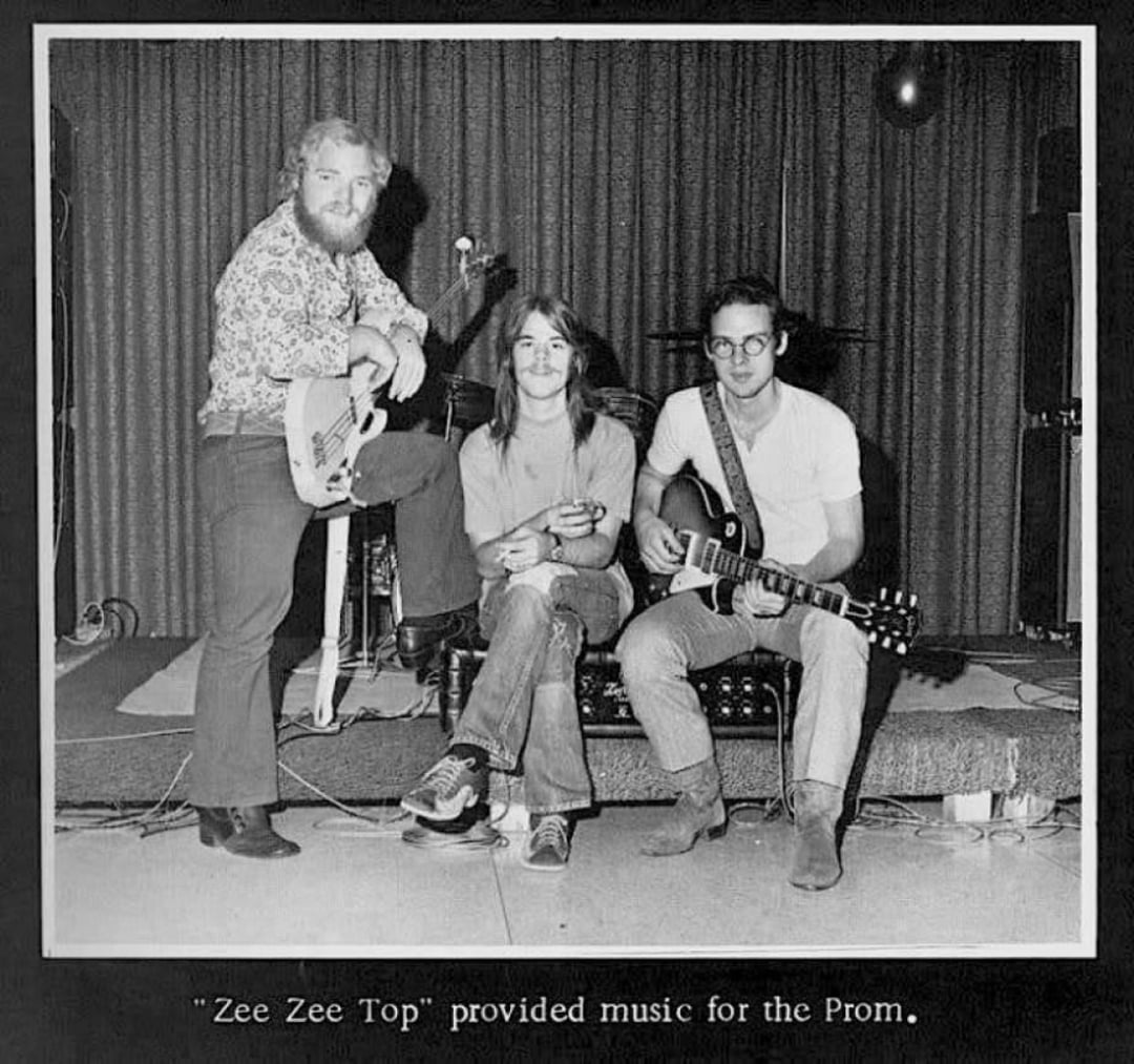 Three young men with guitars pose on a small stage in front of a curtain. Two are seated, one is standing with a foot on an amp. A drum set is partially visible behind them. Caption reads: "'Zee Zee Top' provided music for the Prom.