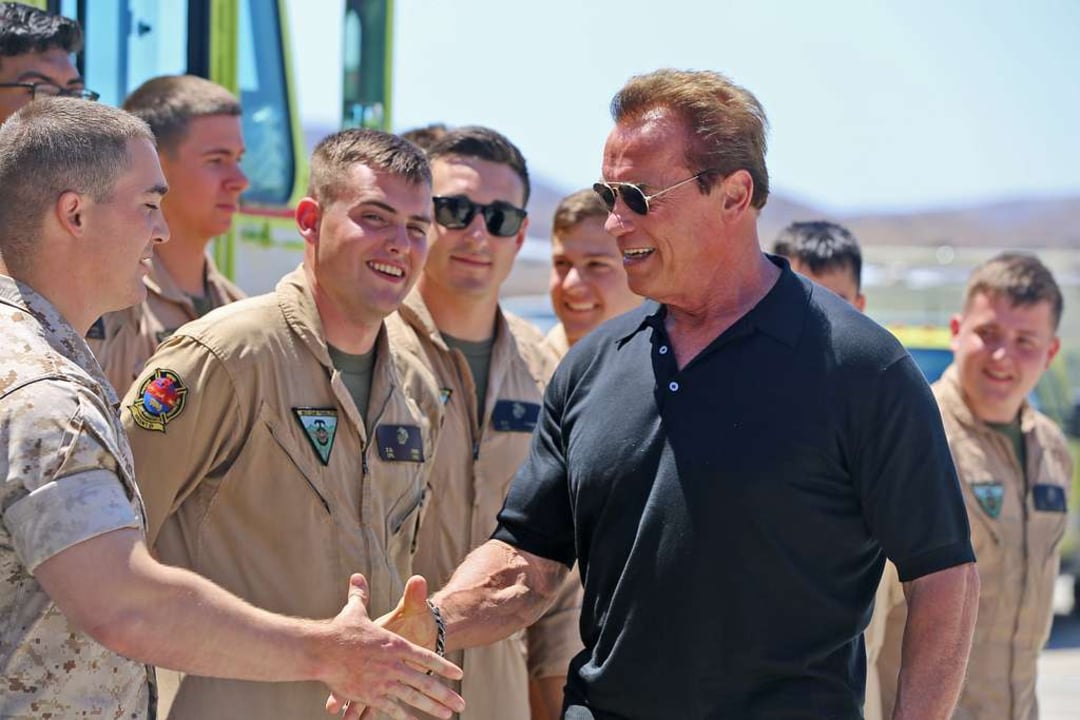 A man in a black shirt smiles and shakes hands with a uniformed service member, surrounded by several other smiling service members outdoors on a sunny day.