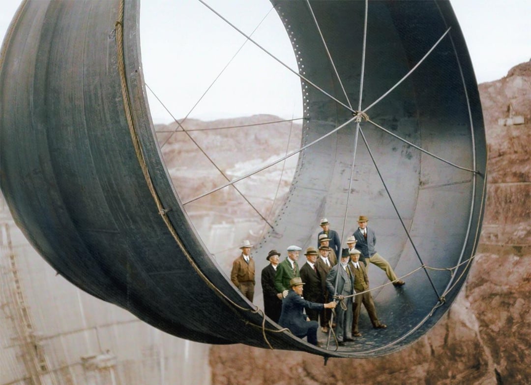 A group of men in 1930s attire stand inside a large steel pipe suspended high above the ground at the Hoover Dam construction site, with rocky terrain visible in the background.