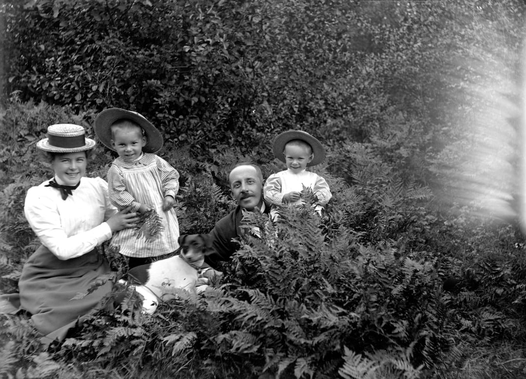 A black and white photo of a family with three children and a dog sitting among ferns outdoors. The adults and children are wearing vintage clothing and hats, smiling at the camera, surrounded by dense foliage.
