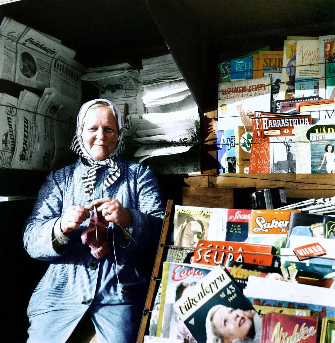 An elderly woman in a headscarf sits knitting beside a kiosk filled with various colorful newspapers and magazines, some in Finnish, with stacks of papers behind her.