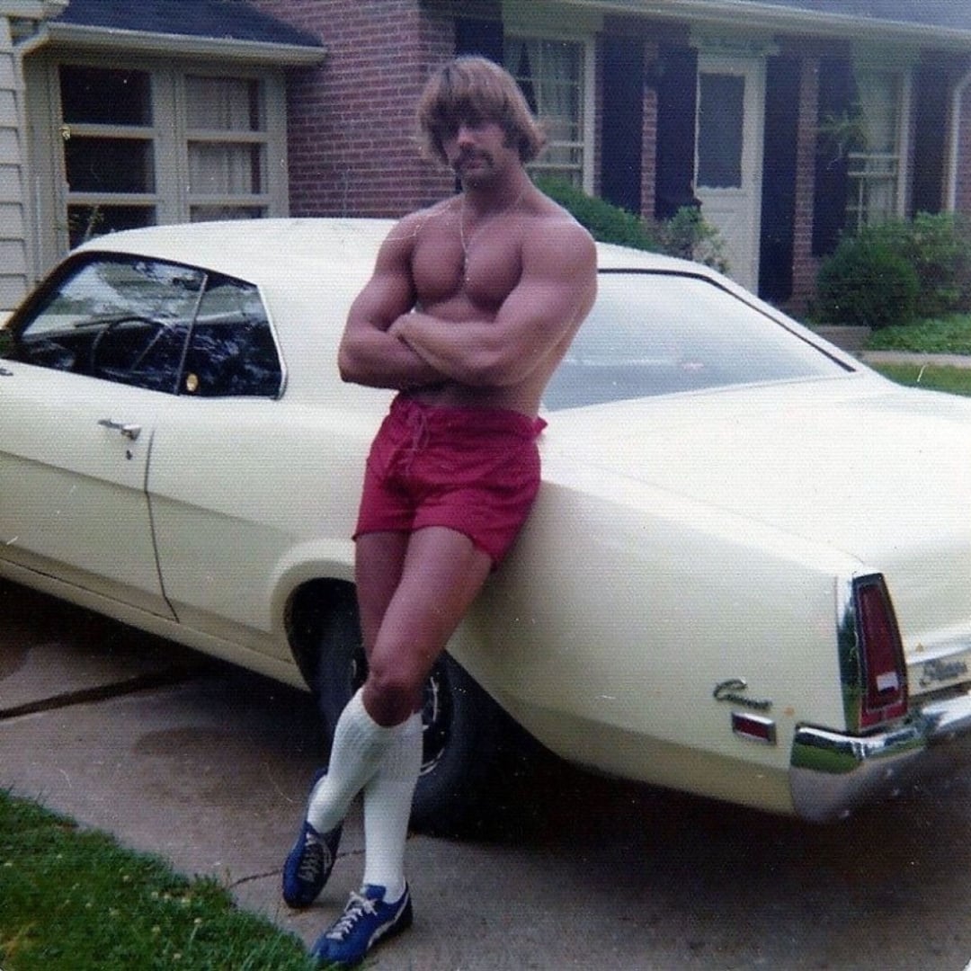A muscular man with a mustache, wearing red shorts, long white socks, and blue sneakers, stands shirtless with arms crossed, leaning against a vintage cream-colored car parked in a driveway.