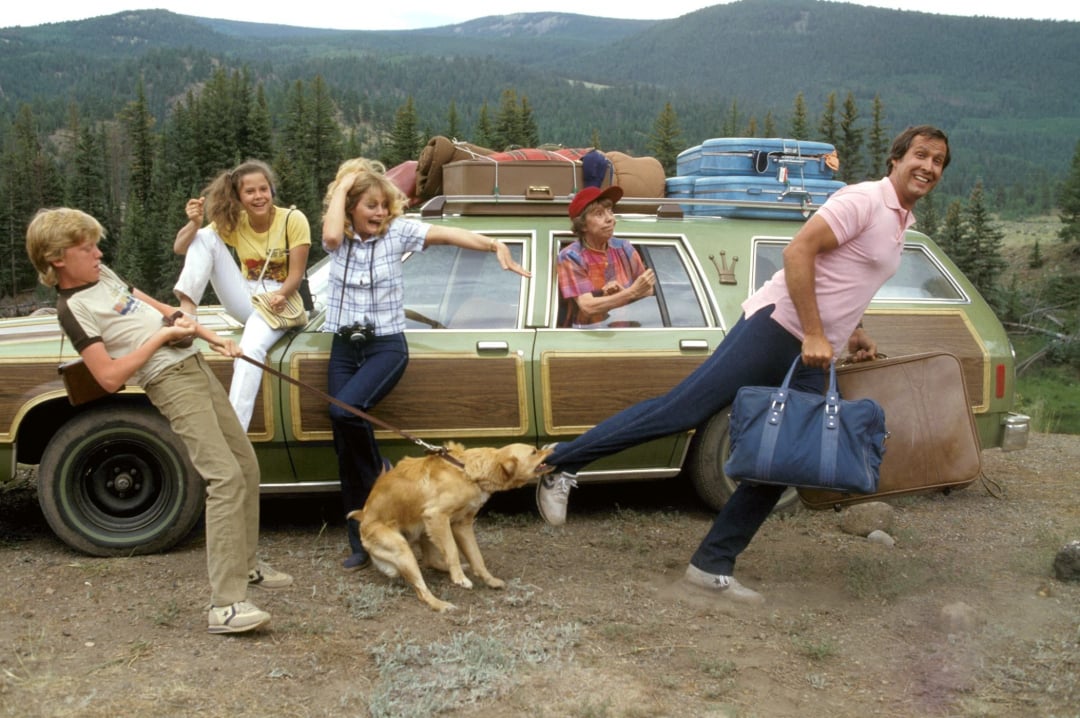 A family stands beside a station wagon packed with luggage in a scenic mountain area. A dog tugs at the man’s leg while the others laugh and react excitedly. Forested hills and mountains are visible in the background.