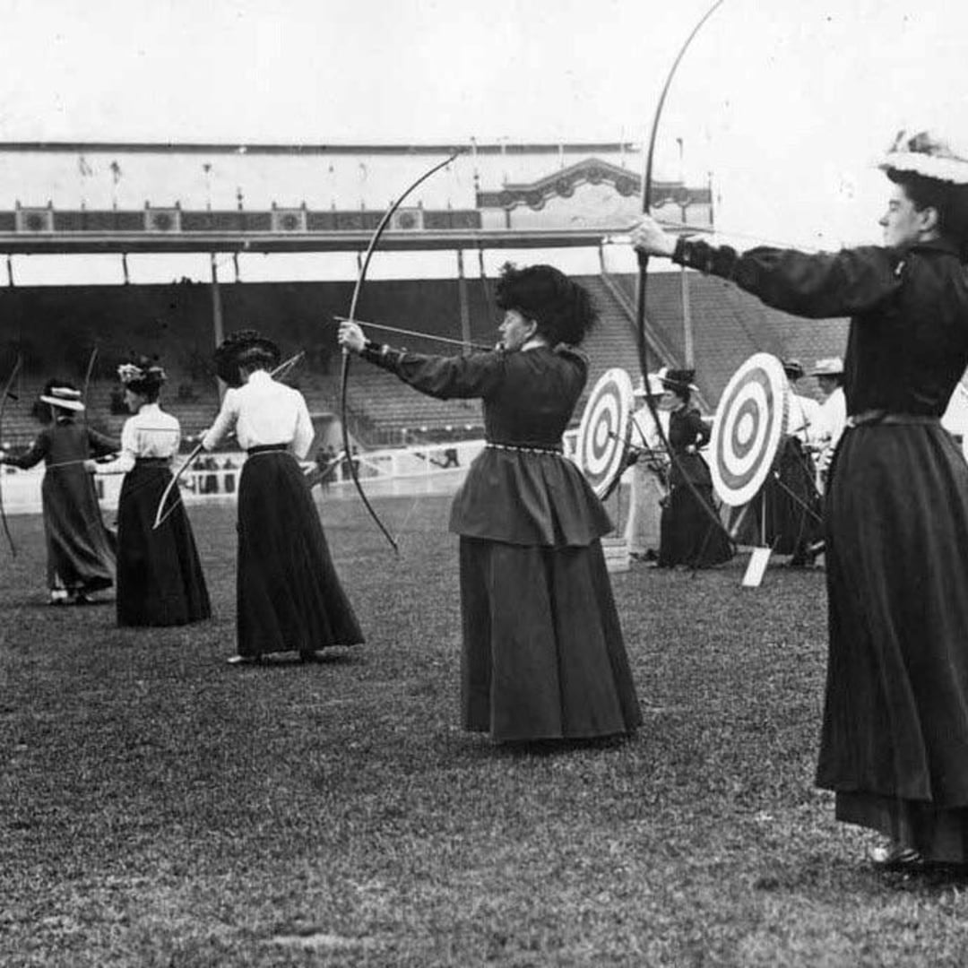Women in long skirts and hats participate in an archery competition on a grass field, aiming at round targets; a stadium with empty seats is visible in the background.