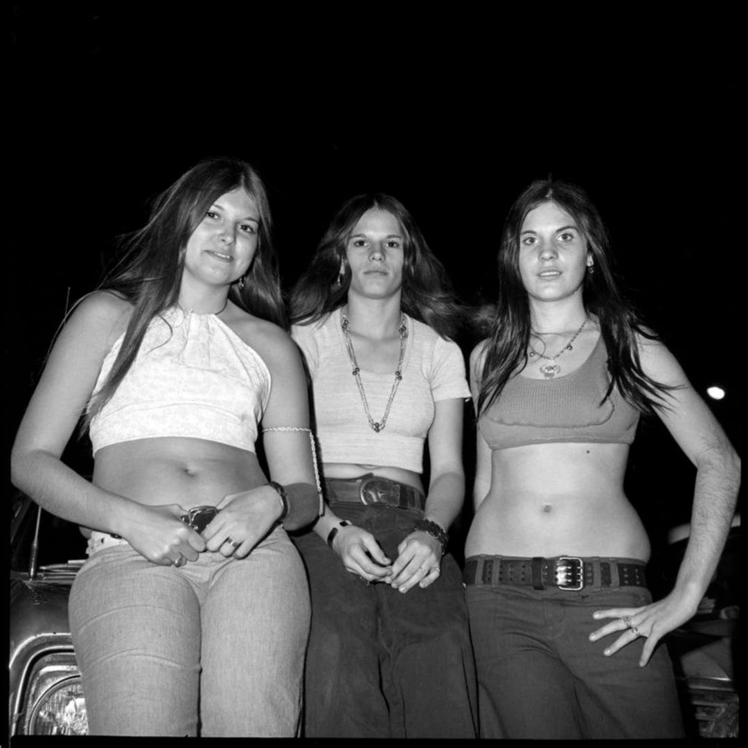 Three young women with long hair pose together at night, wearing crop tops and jeans with belts, standing and sitting on a car against a dark background.