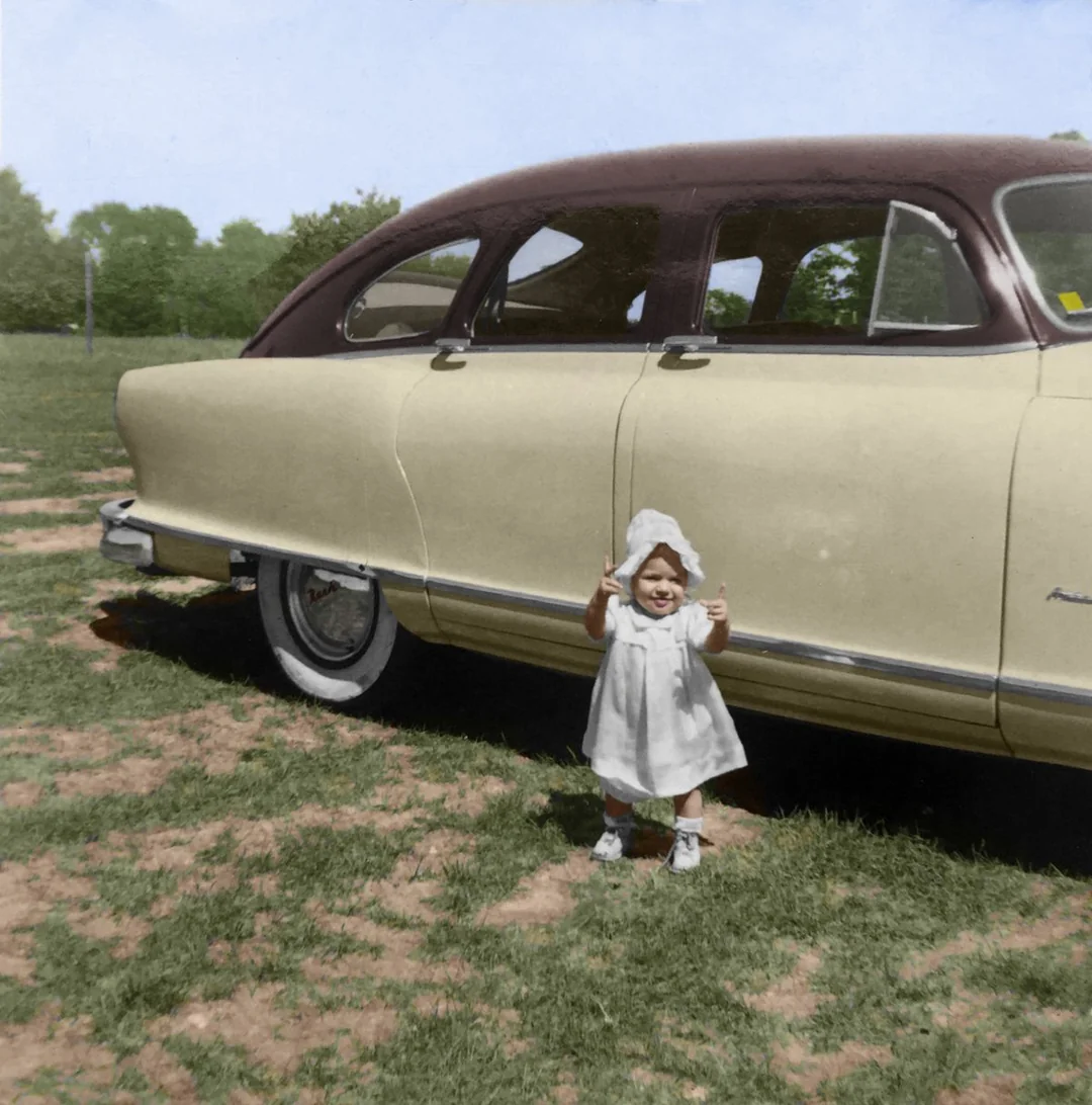 A young child in a white dress and bonnet stands on grass, smiling and giving two thumbs up in front of a vintage beige and brown car parked outdoors.