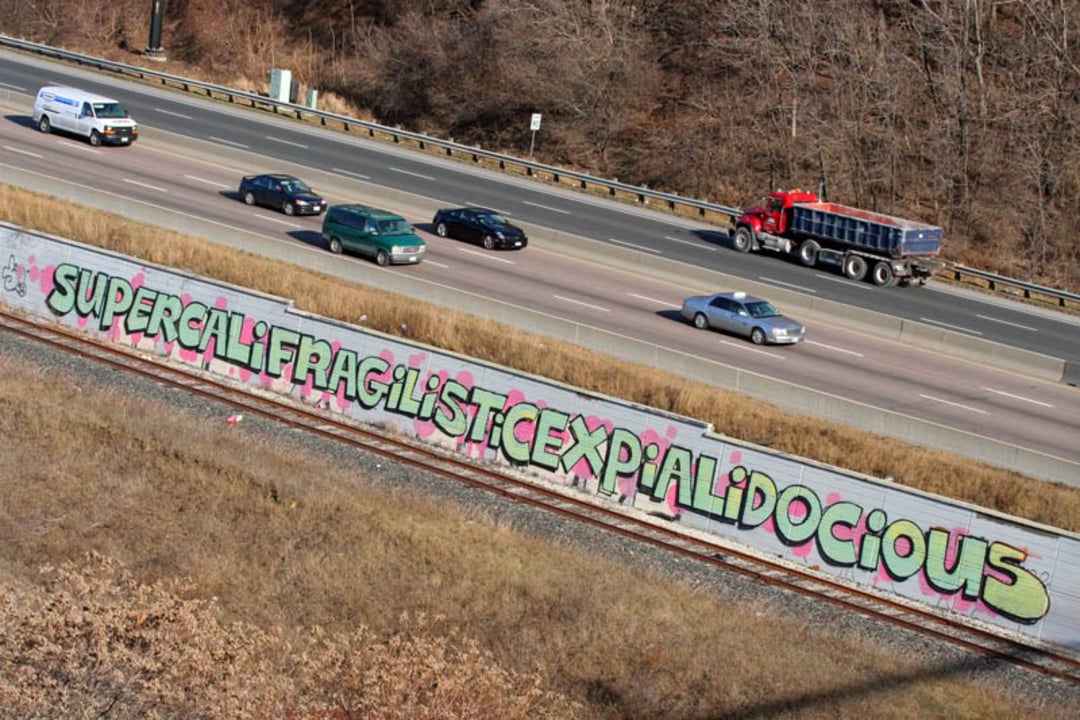 A long graffiti mural along a highway wall spells out "SUPERCALIFRAGILISTICEXPIALIDOCIOUS" in colorful, bold letters as cars and a truck drive by on the adjacent highway.