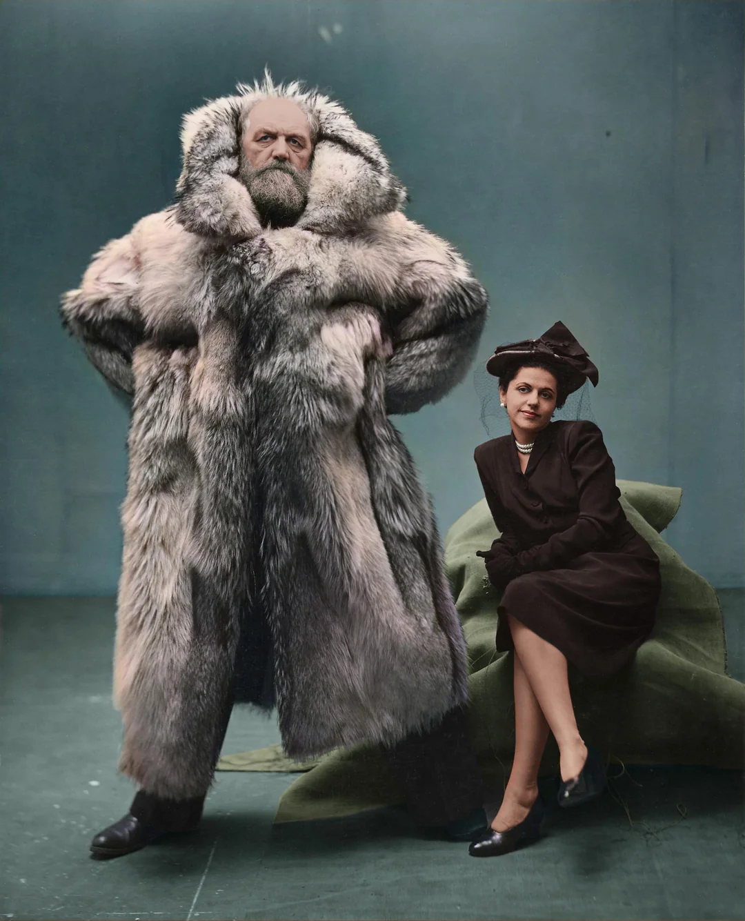 A man stands confidently in a large, luxurious fur coat with his hands on his hips, while a woman in a dark dress and hat sits on a green cloth, looking at the camera. The background is plain and blue-green.
