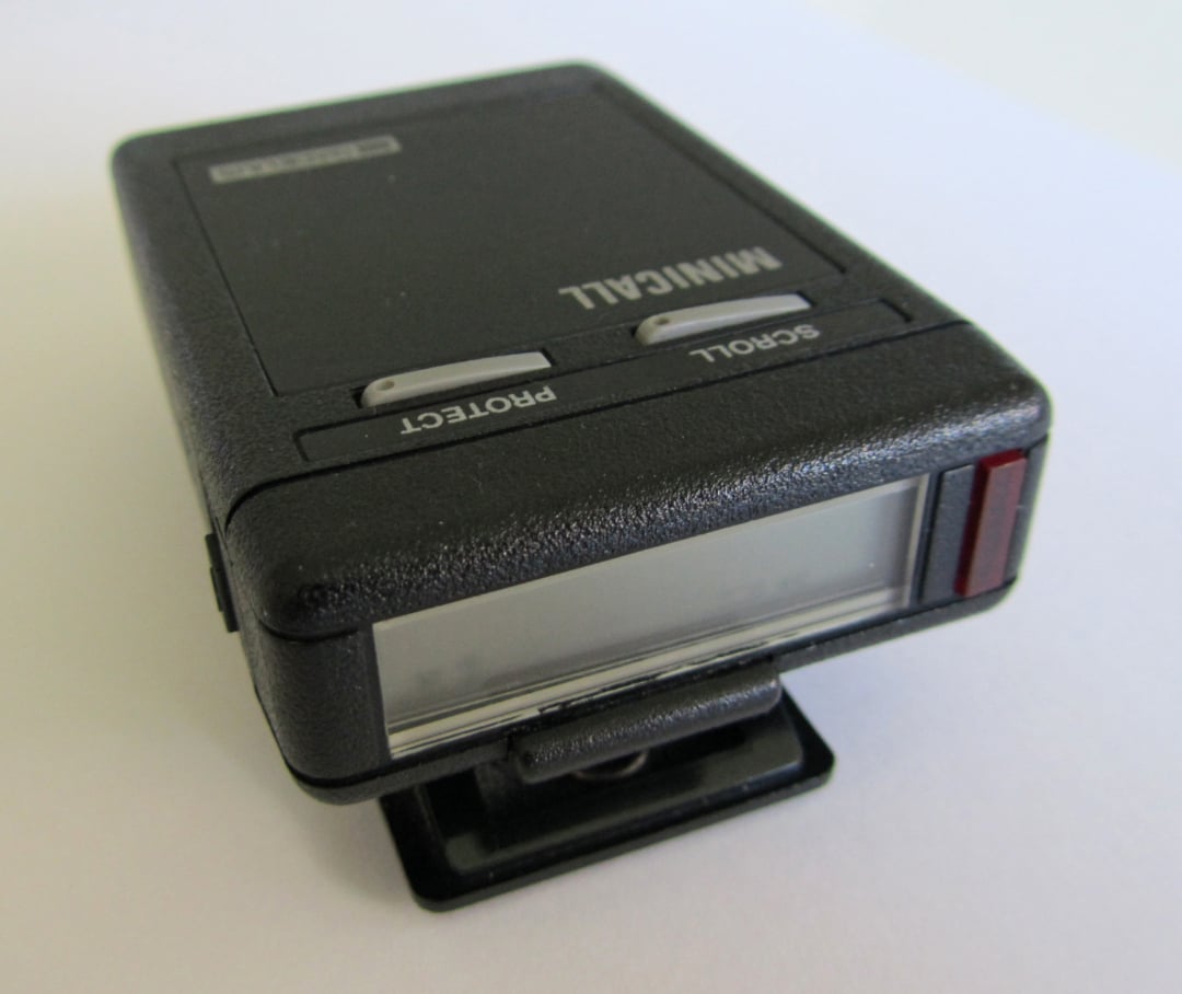 A close-up photo of a vintage black pager with a small display screen, labeled "MINICALL," and buttons for "PROTECT" and "SCROLL." The pager is resting on a white surface.