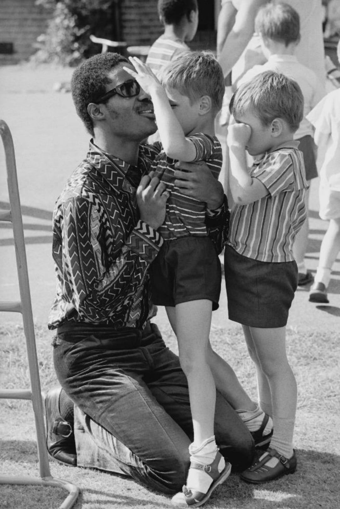 A man wearing sunglasses kneels on grass, smiling as two young boys in striped shirts touch his face. More children are visible in the background outdoors on a sunny day.